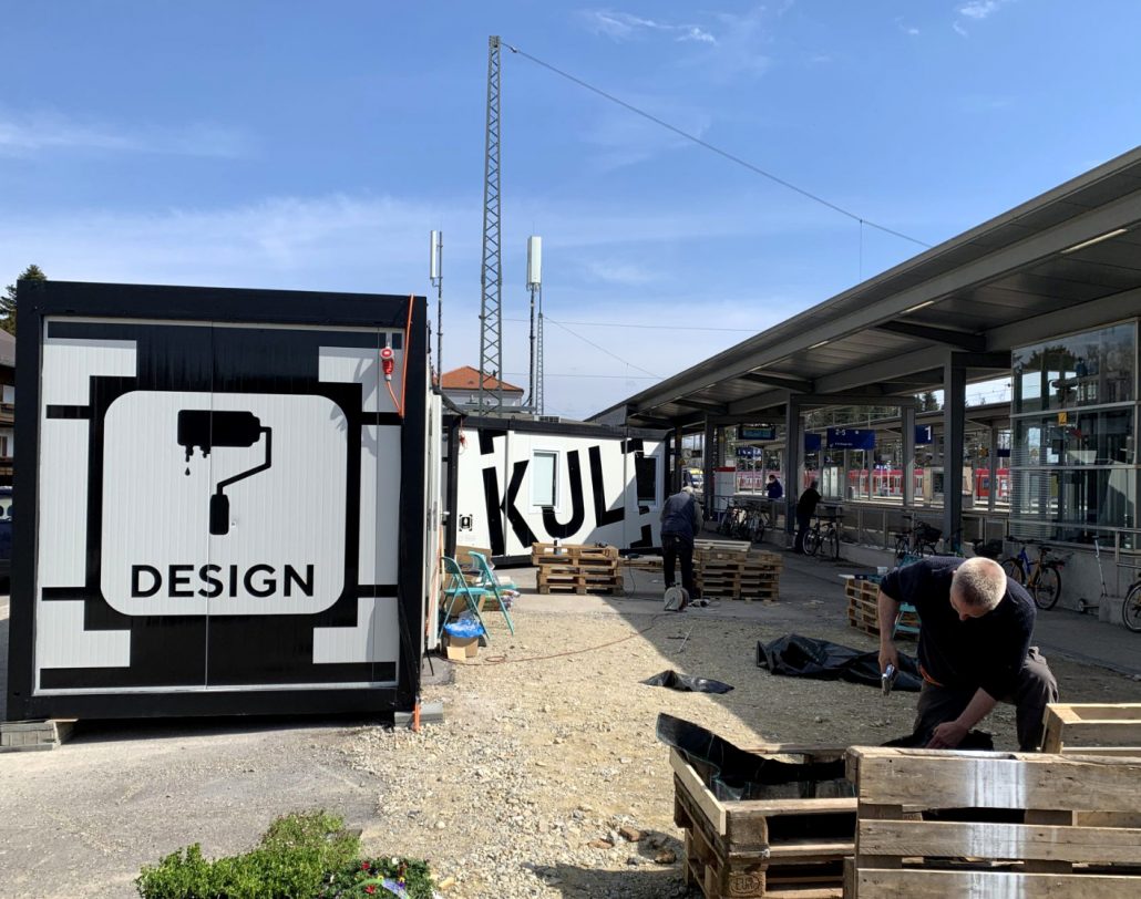 zwei Männer, die Paletten zusammenbauen vor einem Gebäude und Container mit der Schrift "Design"