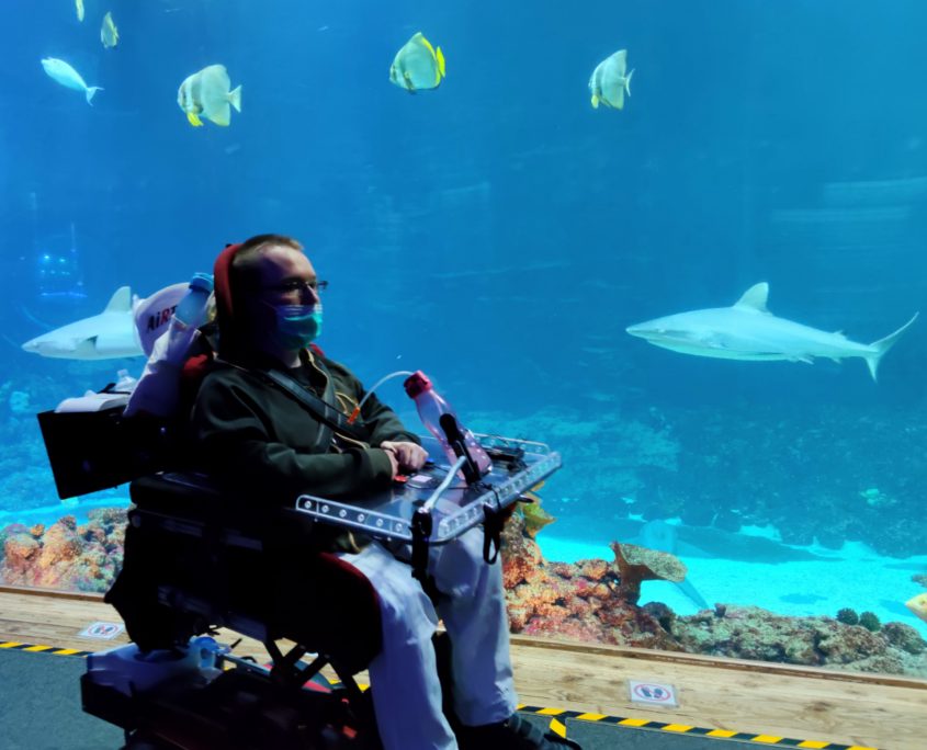 Ein junger Mann im Rollstuhl fährt an einem deckenhohen Aquarium vorbei.