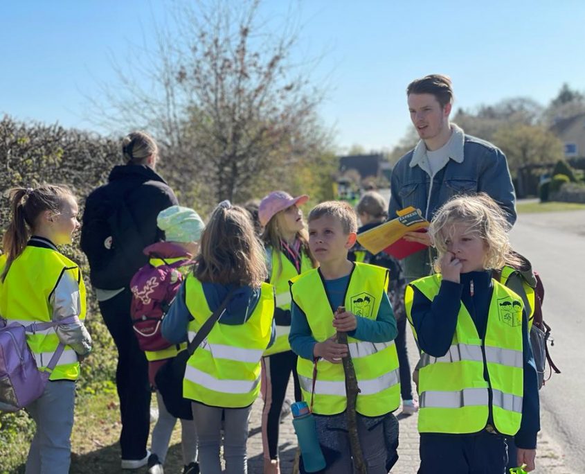 Eine Gruppe Kinder mit Warnwesten sind mit einem Mann zu Fuß unterwegs.