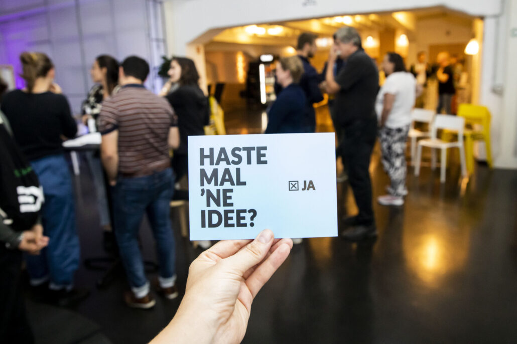 Eine Hand hält eine Postkarte mit dem Text "Haste mal 'ne Idee?". Im Hintergrund mehrere Personen im Gespräch