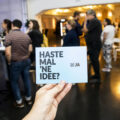 Eine Hand hält eine Postkarte mit dem Text "Haste mal 'ne Idee?". Im Hintergrund mehrere Personen im Gespräch