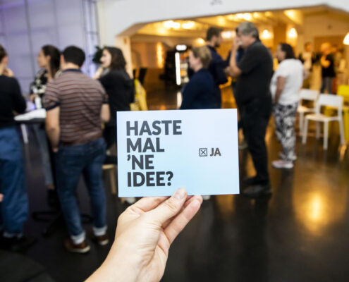 Eine Hand hält eine Postkarte mit dem Text "Haste mal 'ne Idee?". Im Hintergrund mehrere Personen im Gespräch