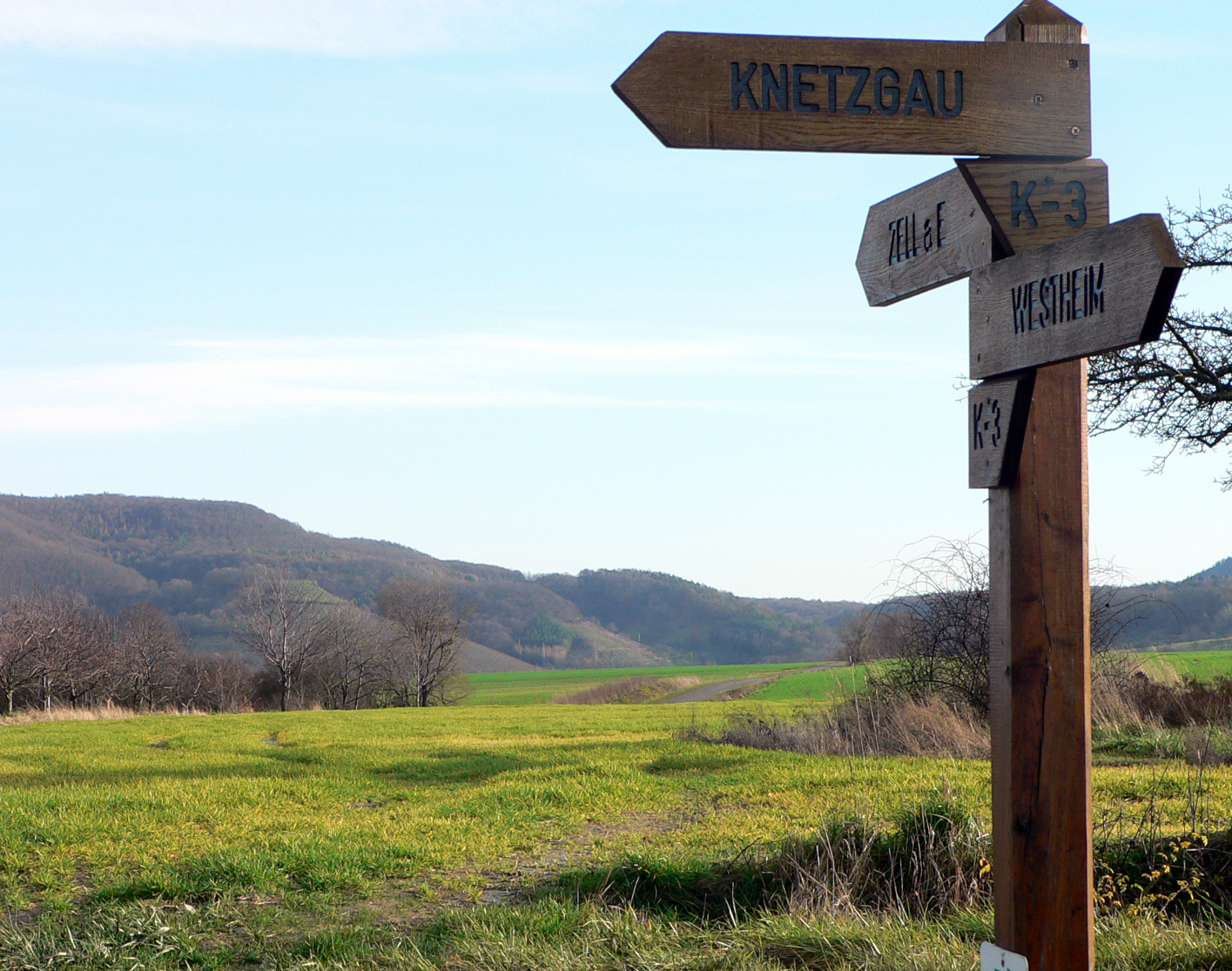 Wegweiser mit dem Schild "Knetzgau" in einer LAndschaft mit Feldern, Sträuchen und Hügeln
