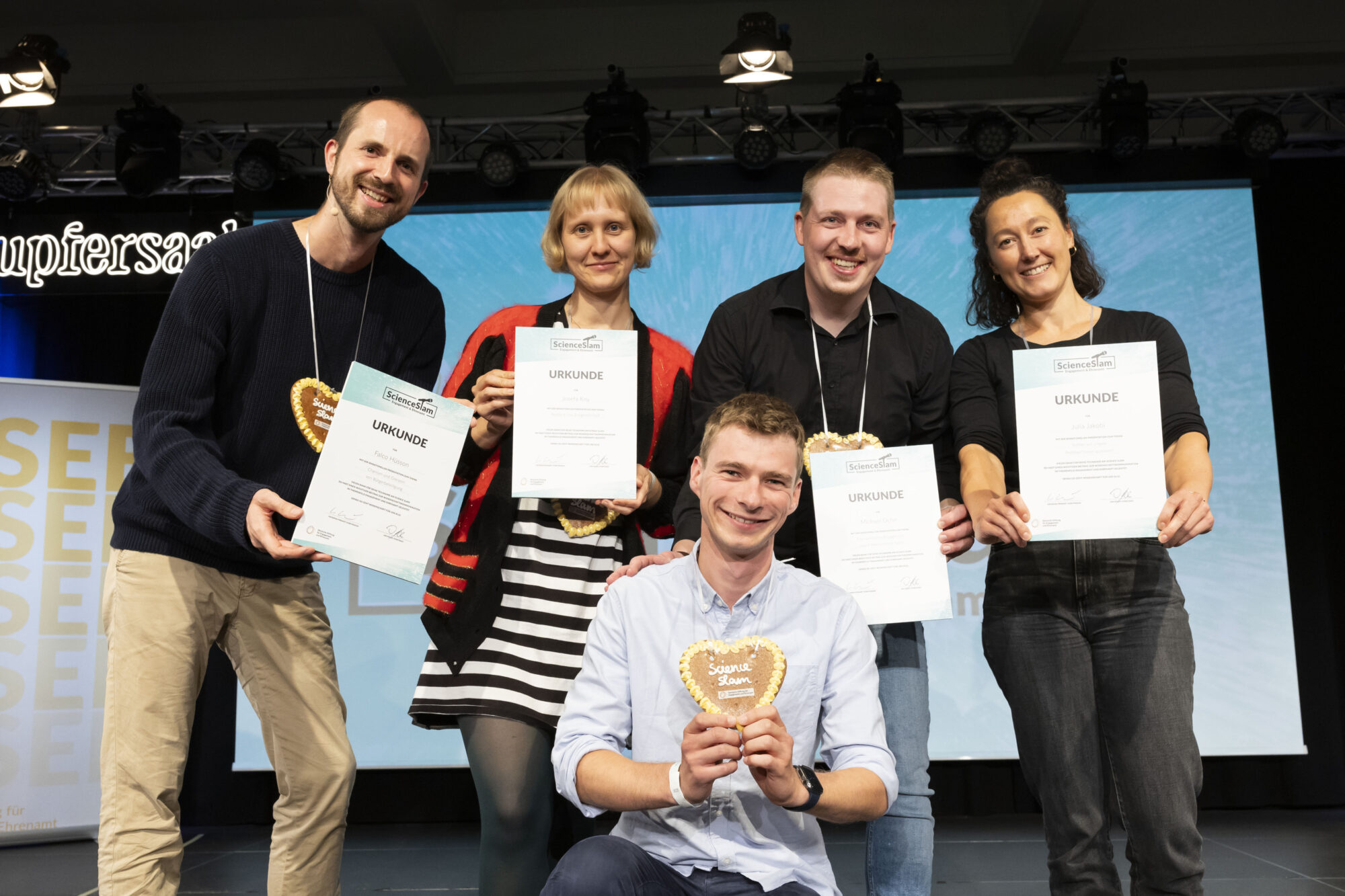 Die Slammies Falco Hüsson, Dr. Josefa Kny, Michael Dehn, Julia Jakobi, und Jonas Deichelmann stehen auf der Bühne und präsentieren ihre Urkunden und ein Lebkuchenherz mit der Aufschrift "Science Slam"