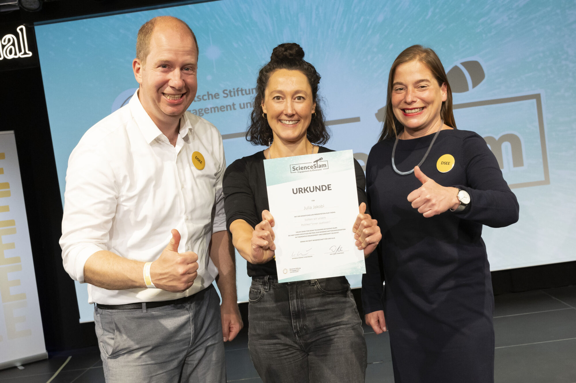 Science-Slam-Gewinnerin Julia Jakobi steht mit DSEE-Vorständin Katarina Peranić und DSEE-Vorstand Jan Holze auf der Bühne und hält ihre Urkunde in die Kamera