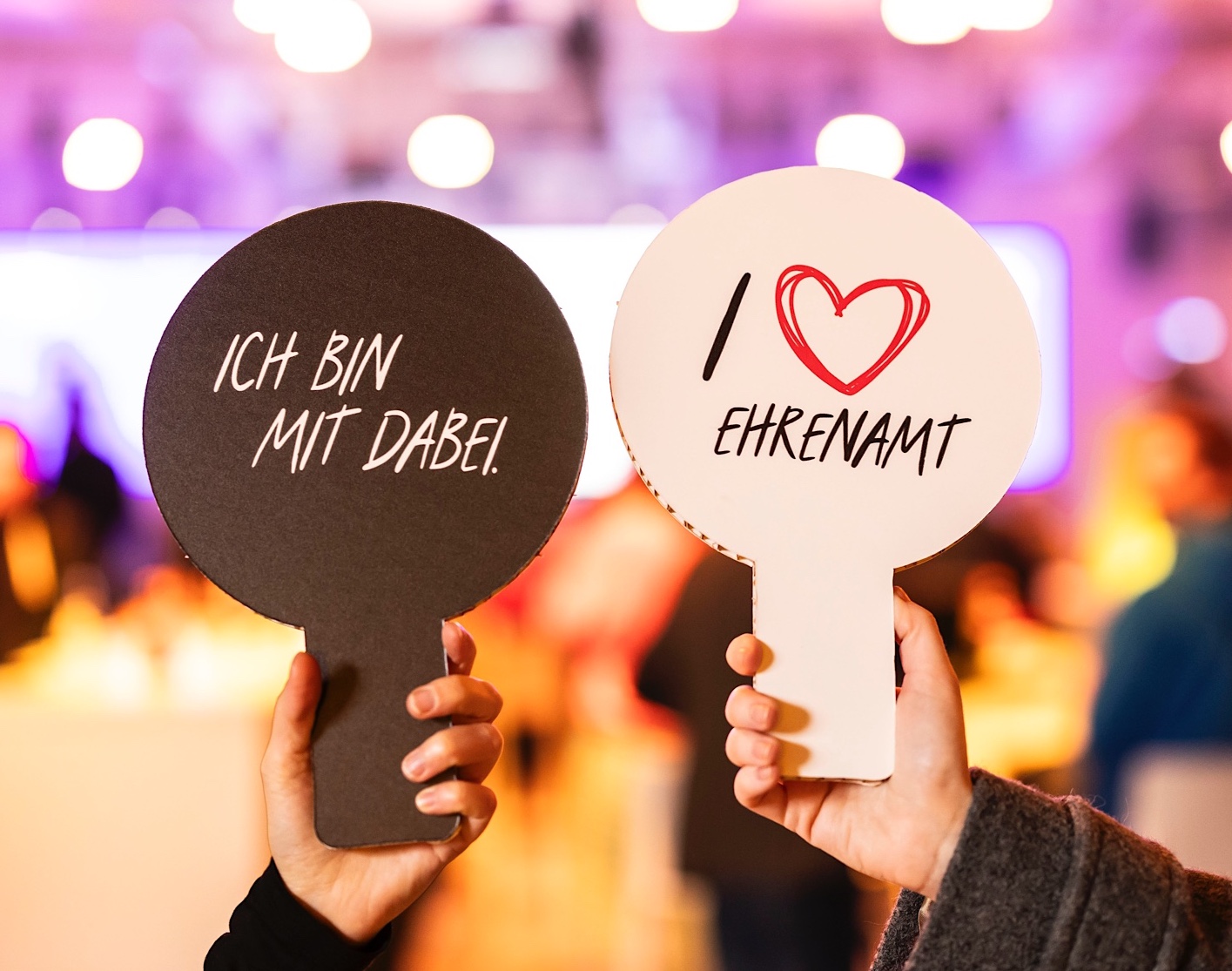 Foto von zwei Händen, die jeweils ein Schild tragen. Auf dem einen steht auf schwarzem Hintergund der Text "Ich bin mit dabei." Auf dem anderen steht auf weißem Hintergrund der Text "Ich liebe Ehrenamt"