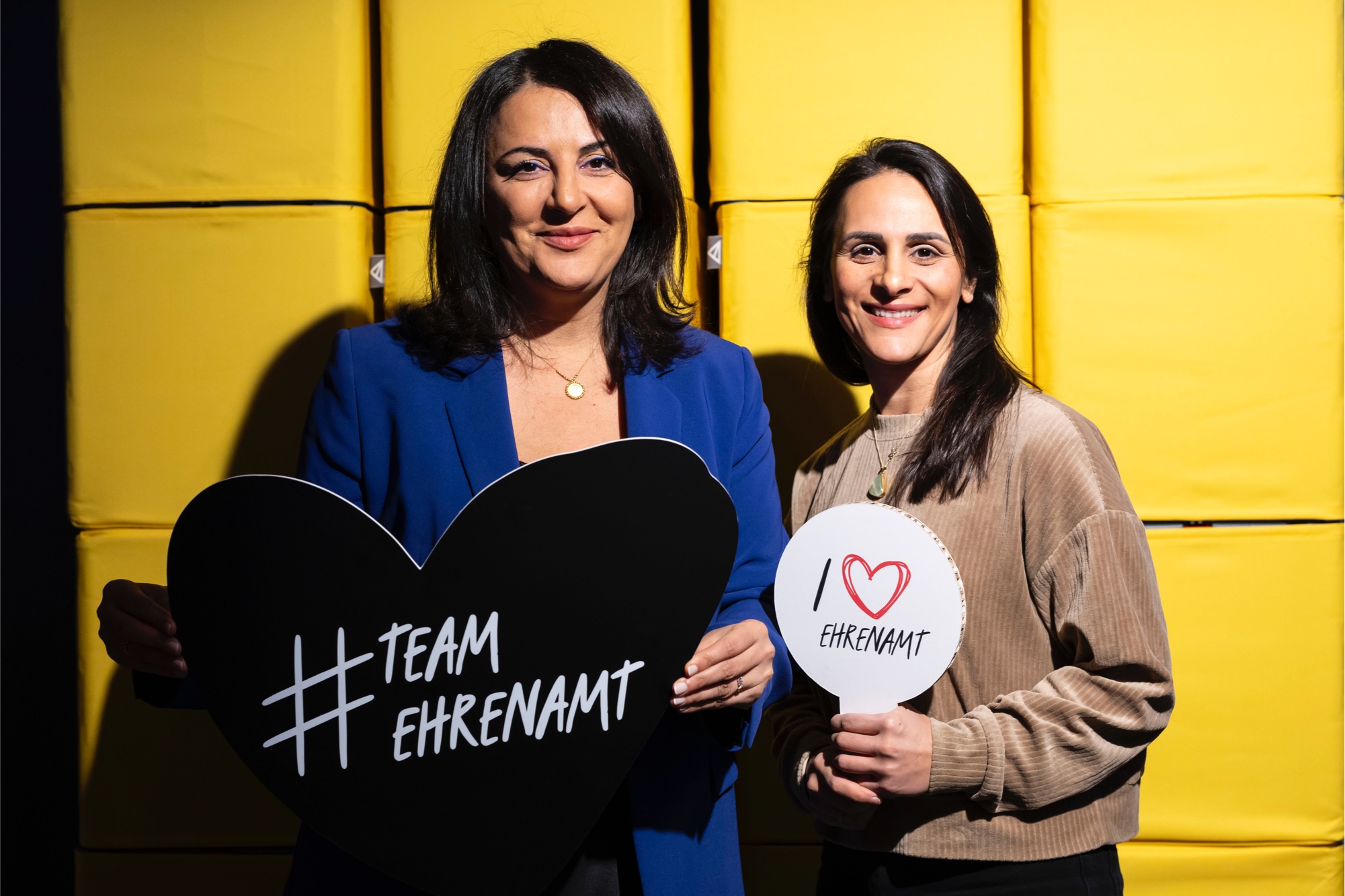 Düzen Tekkal und Tezcan Tekkal halten Schilder mit den Slogans #Team Ehrenamt und I love Ehrenamt in die Kamera