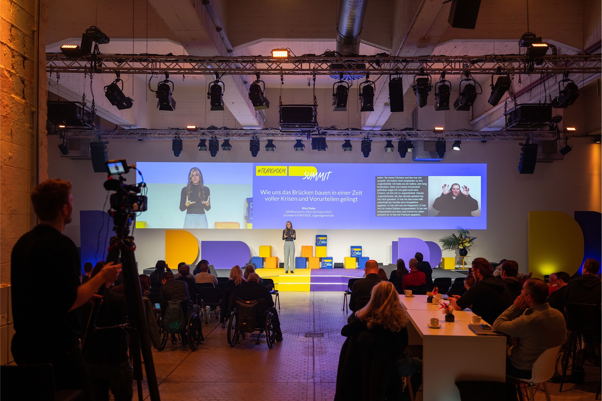 Foto von der Bühne beim transofrm_d Summit. Dort steht Kira Geiss, Miss Germany 2023. Auf der rechten Seite des Bühnenmonitors wird der Vortrag untertitelt und in Gebärden gedolmetscht.