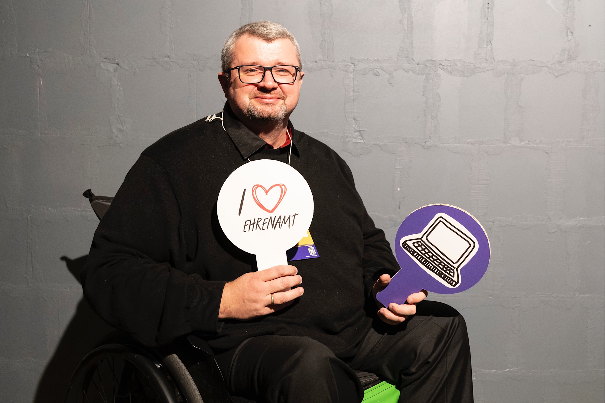 Ein Mann im Rollstuhl hält zwei Schilder in den Händen. Auf dem einen steht "Ich liebe Ehrenamt". Auf dem anderen ist ein gezeichneter Computer.