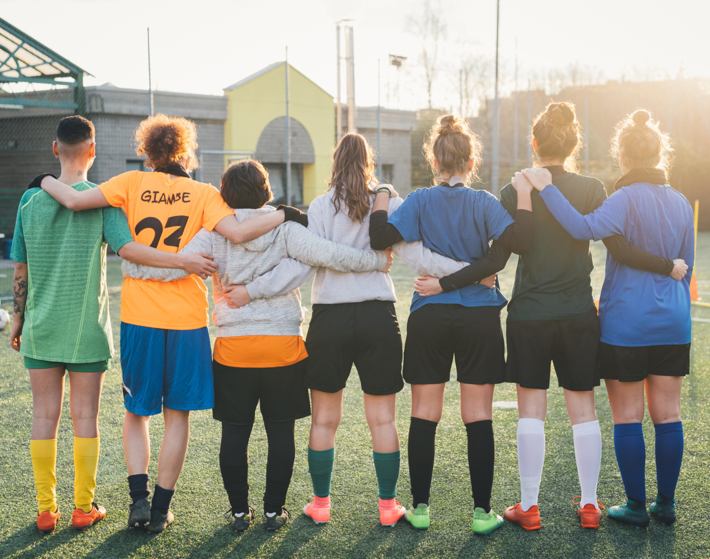 Foto von hinten: Junge Sportlerinnen und Sportler auf dem Sportplatz haben sich untergehakt.