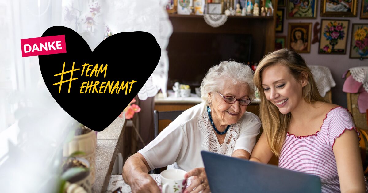 Foto von einer älteren und einer jungen Frau, die nebeneinander sitzen und gemeinsam auf einen Computer schauen. Ein gezeichnetes Herz mit dem Text: Danke #TeamEhrenamt