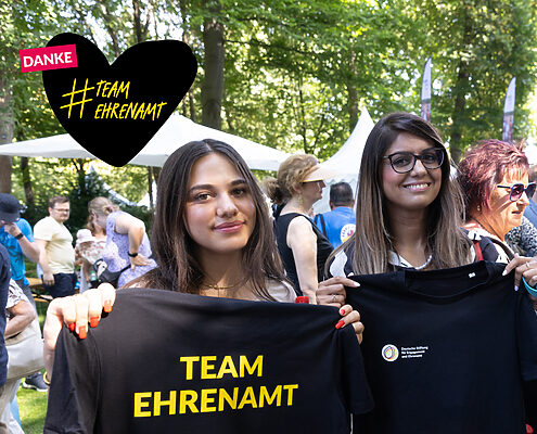 Foto von zwei jungen Frauen, die T-Shirts mit dem Logo der DSEE und dem Schriftzug Team Ehrenamt in die Hände halten. Gezeichnetes Herz mit dem Text Danke #TeamEhrenamt