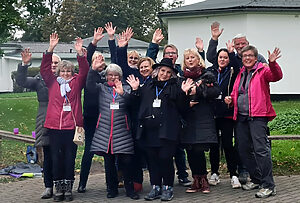 Eine Gruppe von 12 Menschen verschiedenen Alters, die in die Kamera winken und lächeln.