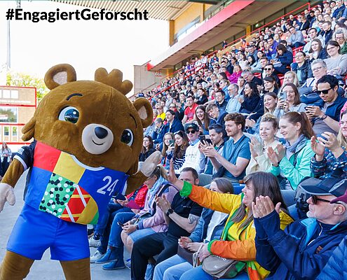 Freiwillig Engagierte interagieren auf der Tribüne eines Stadions sitzend mit dem lebenden Maskotchen der UEFA 2024, einem Bären mit Krone.