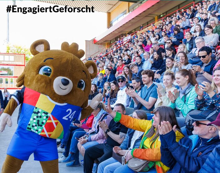 Freiwillig Engagierte interagieren auf der Tribüne eines Stadions sitzend mit dem lebenden Maskotchen der UEFA 2024, einem Bären mit Krone.