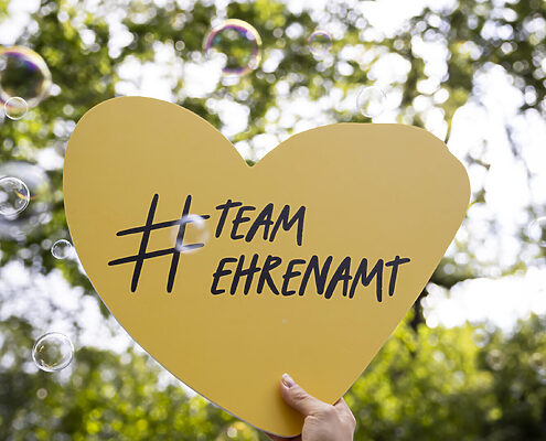 Hochgehaltenes Herz mit der Beschriftung "# Team Ehrenamt" (Hashtag und Wörter zusammengeschrieben). Im Hintergrund Seifenblasen.