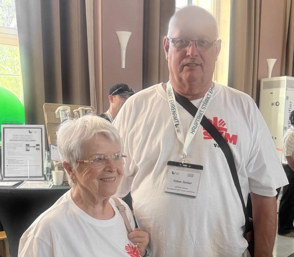 Zwei Personen in weißen Vereins-T-Shirts stehen gemeinsam bei einer Veranstaltung in einem festlich dekorierten Saal.