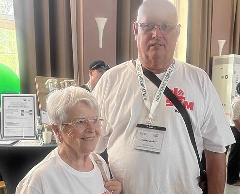 Zwei Personen in weißen Vereins-T-Shirts stehen gemeinsam bei einer Veranstaltung in einem festlich dekorierten Saal.