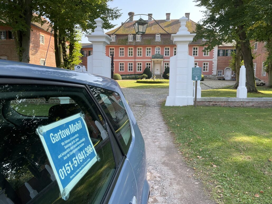 Ein Fahrzeug mit blauem GartowMobil-Schild an der Seitenscheibe steht auf einem Kiesweg vor dem historischen Gartower Schloss, einem rosafarbenen Barockgebäude mit weißen Säulen am Eingang, umgeben von grünen Rasenflächen und alten Bäumen.