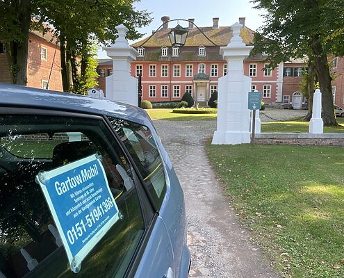 Ein Fahrzeug mit blauem GartowMobil-Schild an der Seitenscheibe steht auf einem Kiesweg vor dem historischen Gartower Schloss, einem rosafarbenen Barockgebäude mit weißen Säulen am Eingang, umgeben von grünen Rasenflächen und alten Bäumen.