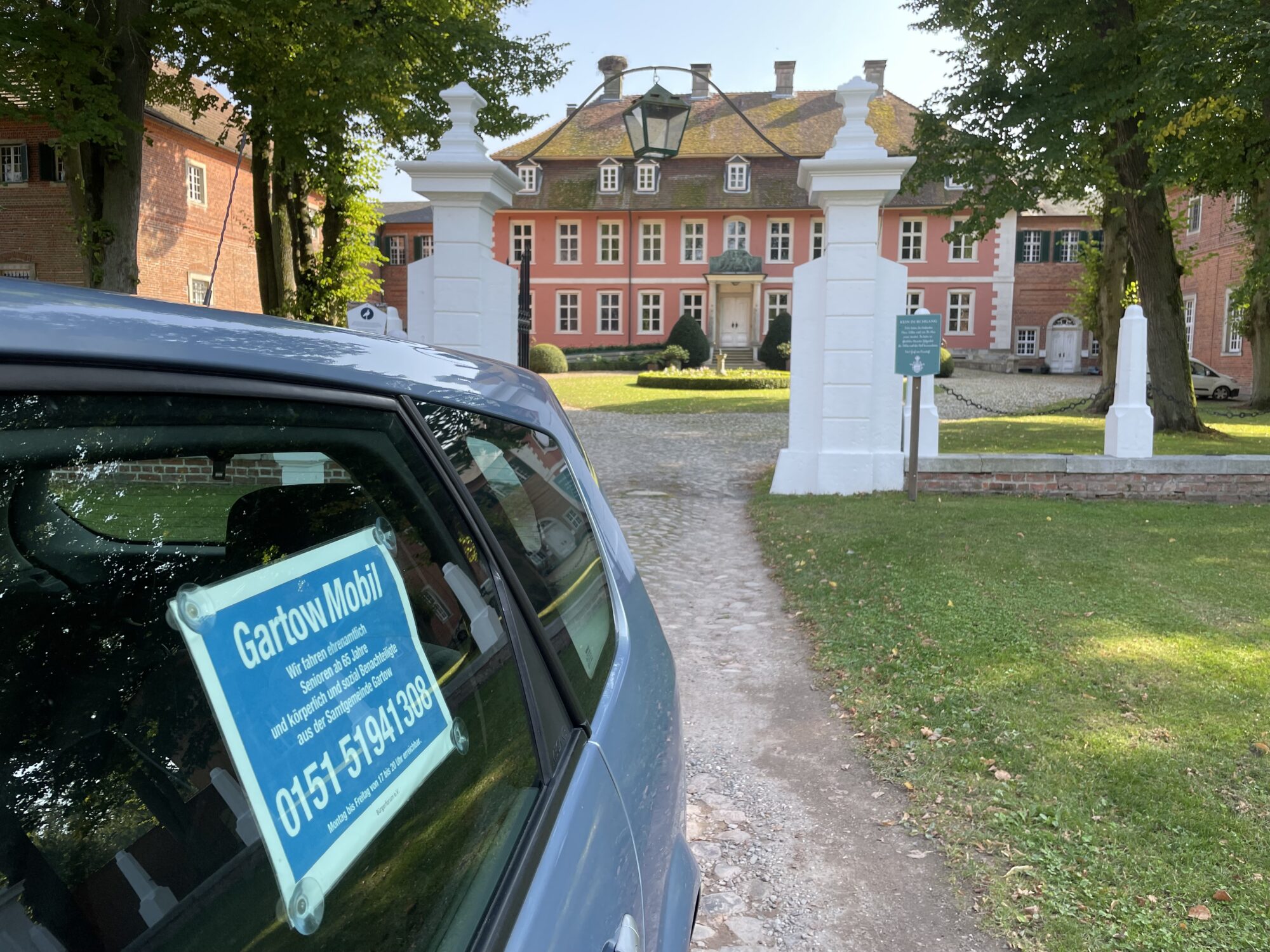 Ein Fahrzeug mit blauem GartowMobil-Schild an der Seitenscheibe steht auf einem Kiesweg vor dem historischen Gartower Schloss, einem rosafarbenen Barockgebäude mit weißen Säulen am Eingang, umgeben von grünen Rasenflächen und alten Bäumen.