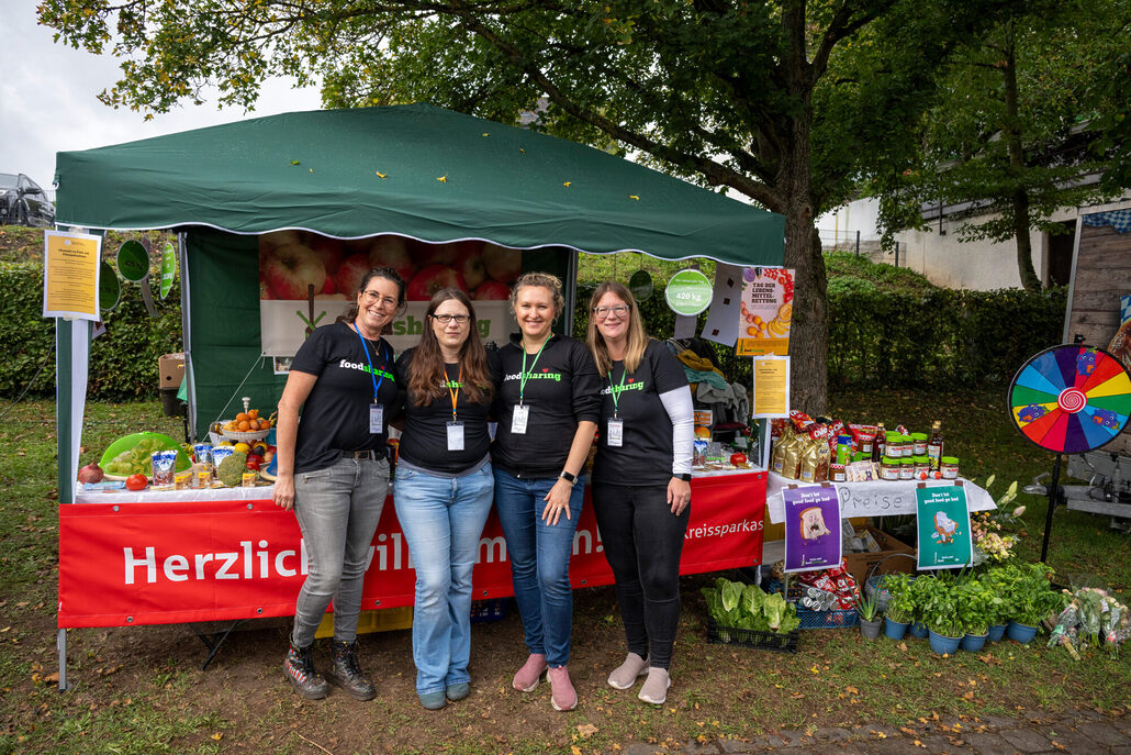 Vier Personen stehen vor einem Info-Stand mit Lebensmitteln und Pflanzen auf einem Fest im Freien. Ein Glücksrad steht am Rand des Stands.