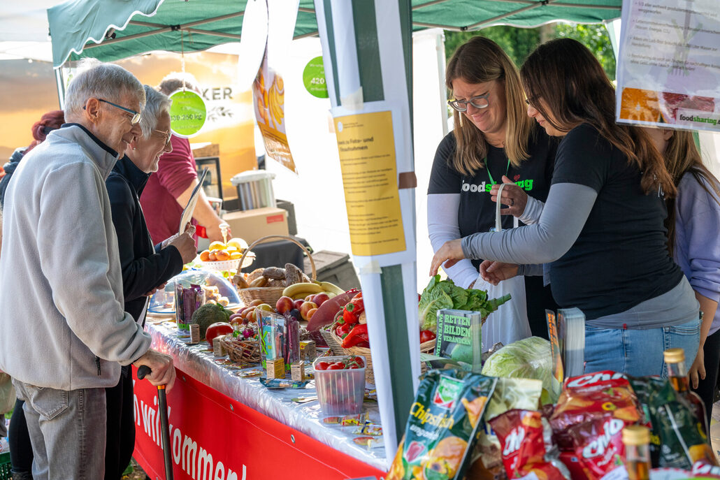 Menschen stehen an einem Marktstand mit Obst, Gemüse und Lebensmitteln und informieren sich bei ehrenamtlichen Helferinnen von foodsharing.