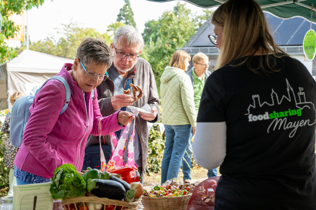 Besucher:innen informieren sich an einem foodsharing-Stand und nehmen Lebensmittel aus einem Korb entgegen. Im Vordergrund ist eine Person mit „foodsharing Mayen“-T-Shirt zu sehen.