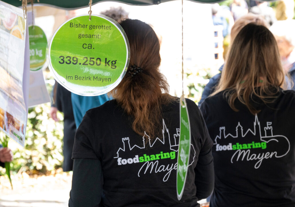 Zwei Personen mit „foodsharing Mayen“-T-Shirts stehen unter Schildern, die auf die gerettete Lebensmittelmenge hinweisen. Ein Schild zeigt: "Bisher gerettet: ca. 339.250 kg im Bezirk Mayen."