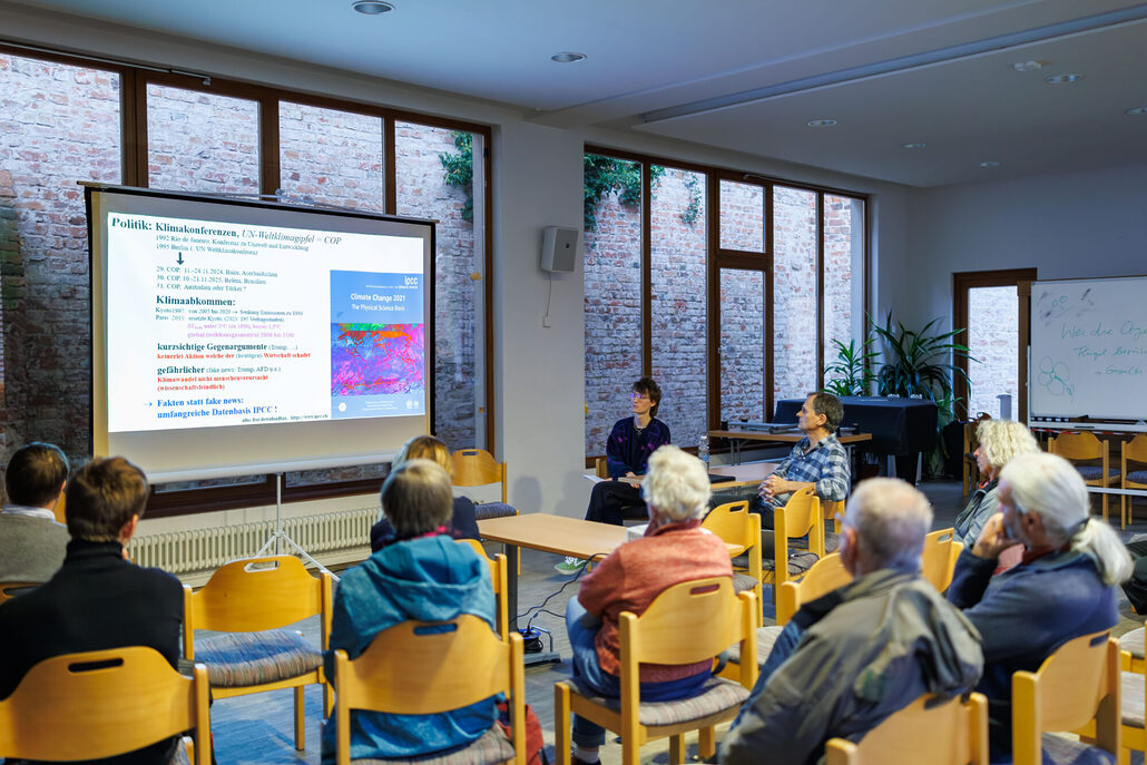 Mehrere Personen sitzen in einem Veranstaltungsraum und verfolgen eine Präsentation zum Thema Klimakonferenzen und Klimawandel, die auf einer großen Leinwand gezeigt wird.