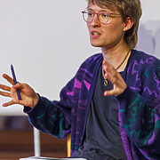 Eine Person sitzt mit Notizbuch und Stift in der Hand und spricht gestikulierend während einer Veranstaltung.